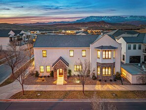 A modern two-story house with a brick entrance and multiple windows, surrounded by a residential area with similar houses and a mountain range in the background. - 5 Suites, Private Pool, 2 Hot Tubs for 19, 2 Fire Pits, 2 Fridges Sleeps 30 (Hurricane)