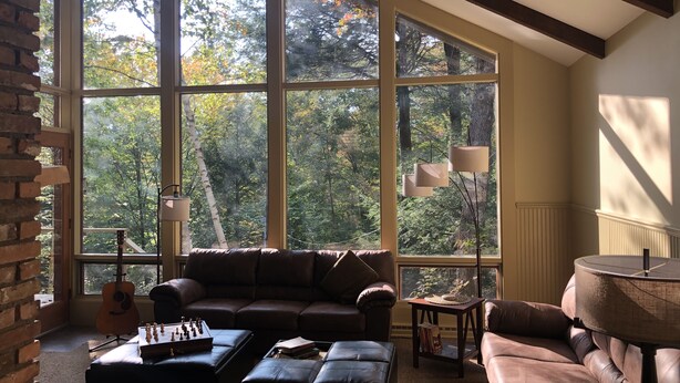A spacious living room with large windows, a ceiling fan, a brown leather sofa, a coffee table with chess, and a lamp. - Green Mountains 4BR House (Peru)