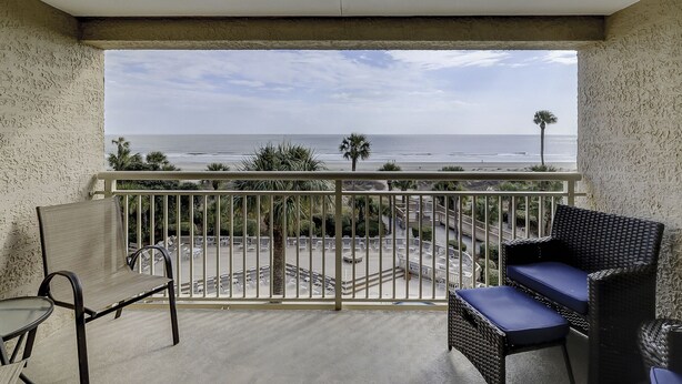 A balcony with a view of the beach and ocean. There are two wicker chairs with cushions.