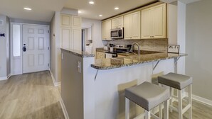 A modern kitchen with white cabinetry, a granite countertop, a microwave, an oven, and a sink. There are two bar stools and a sign indicating Wi-Fi availability.