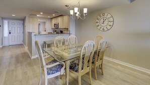 A dining area with a glass table and wooden chairs, a chandelier, and a wall clock.