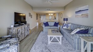 A living room with a sofa, coffee table, and TV. There is a kitchen in the background.