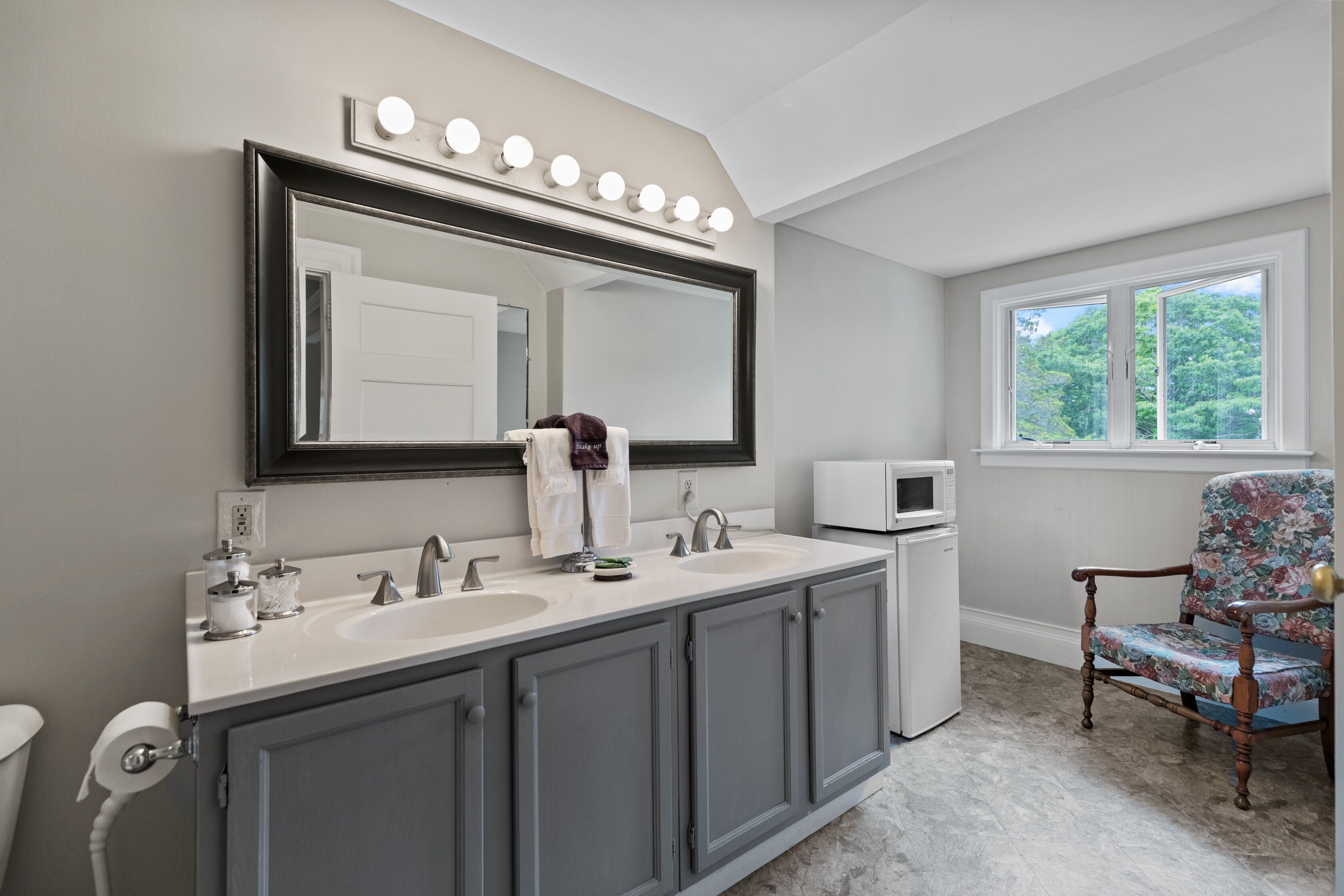 A bathroom with a large mirror, two sinks, a vanity, a microwave, and a chair.