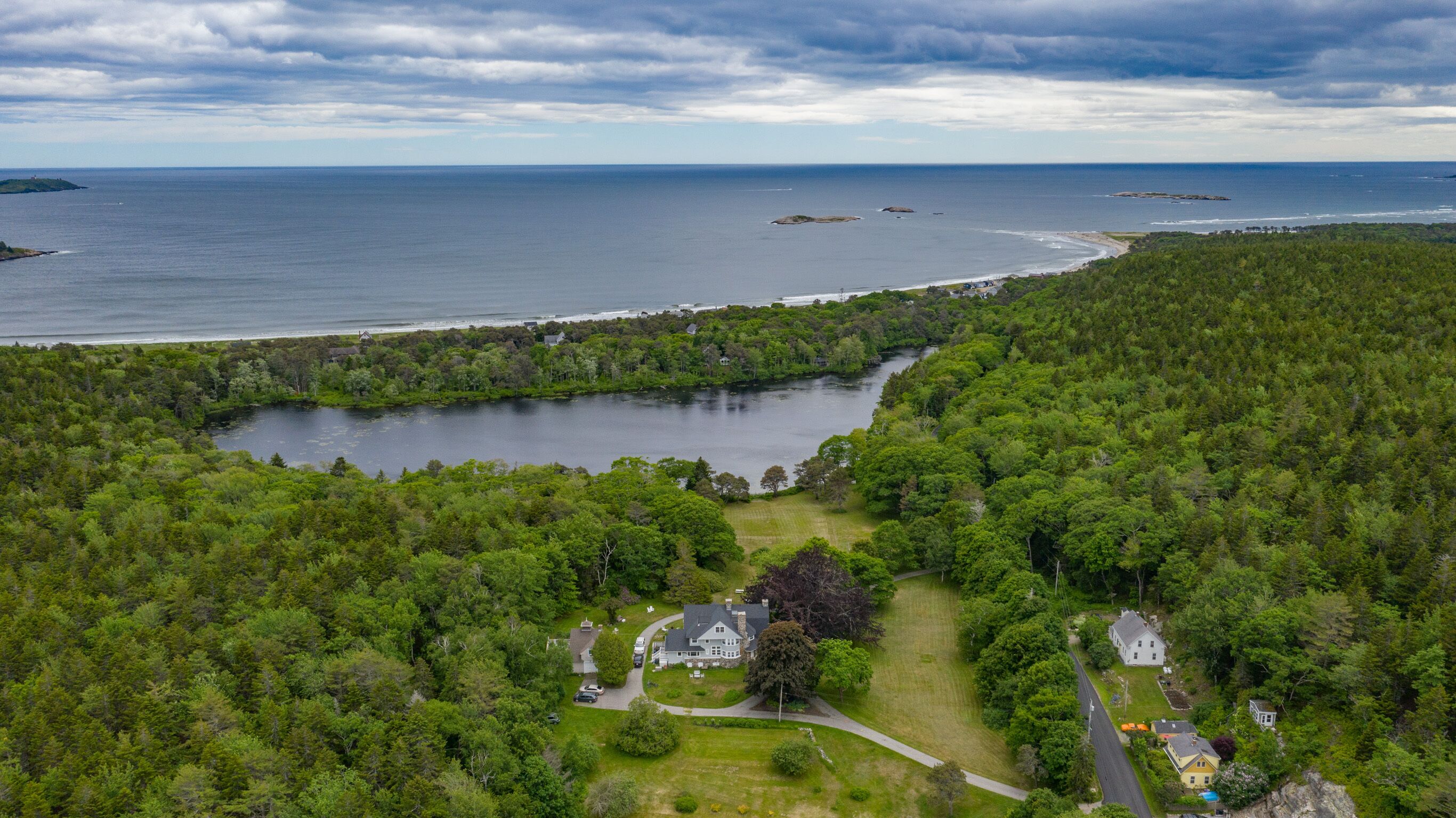 An aerial view of a coastal property with a large lake, dense forest, and a small house with a driveway and parked cars.