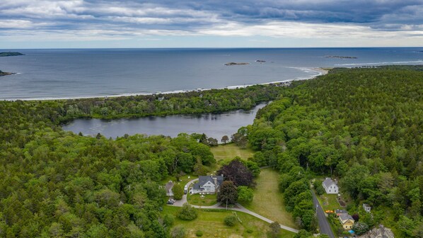 An aerial view of a coastal property with a large lake, dense forest, and a small house with a driveway and parked cars.