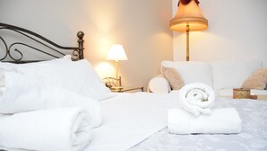 Una cama bien hecha con sábanas blancas, una toalla enrollada, una lámpara de noche y una mesita con una vela.