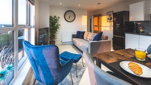 Ein modernes Wohnzimmer mit einer Couch, einem blauen Sessel, einem Esstisch mit Essen und einem Glas Orangensaft, sowie einer Küchenzeile mit Kühlschrank und Schränken.