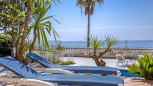 Un espace aménagé au bord de la piscine, avec des chaises longues, un palmier et une vue sur la mer.