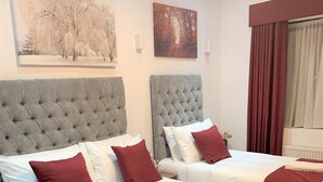A hotel room with two beds, each with a grey tufted headboard, white bedding, and red pillows. There are artwork on the walls and a window with curtains. - Green View Hotel Near Dartford (Dartford)