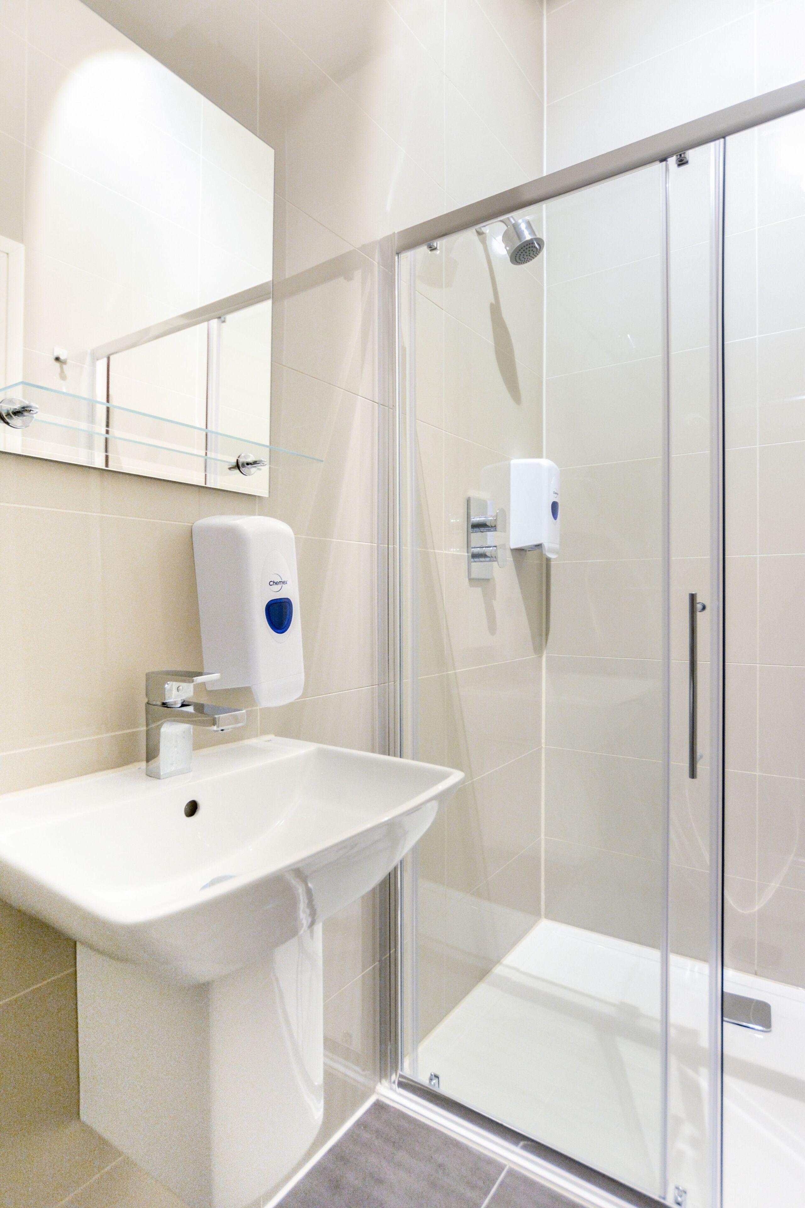A modern bathroom with a glass shower enclosure, a white sink, and a hand soap dispenser.