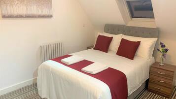 Una cama bien hecha con ropa de cama blanca y roja, un cabecero capitonado gris y una mesita de noche con un jarrón de flores.