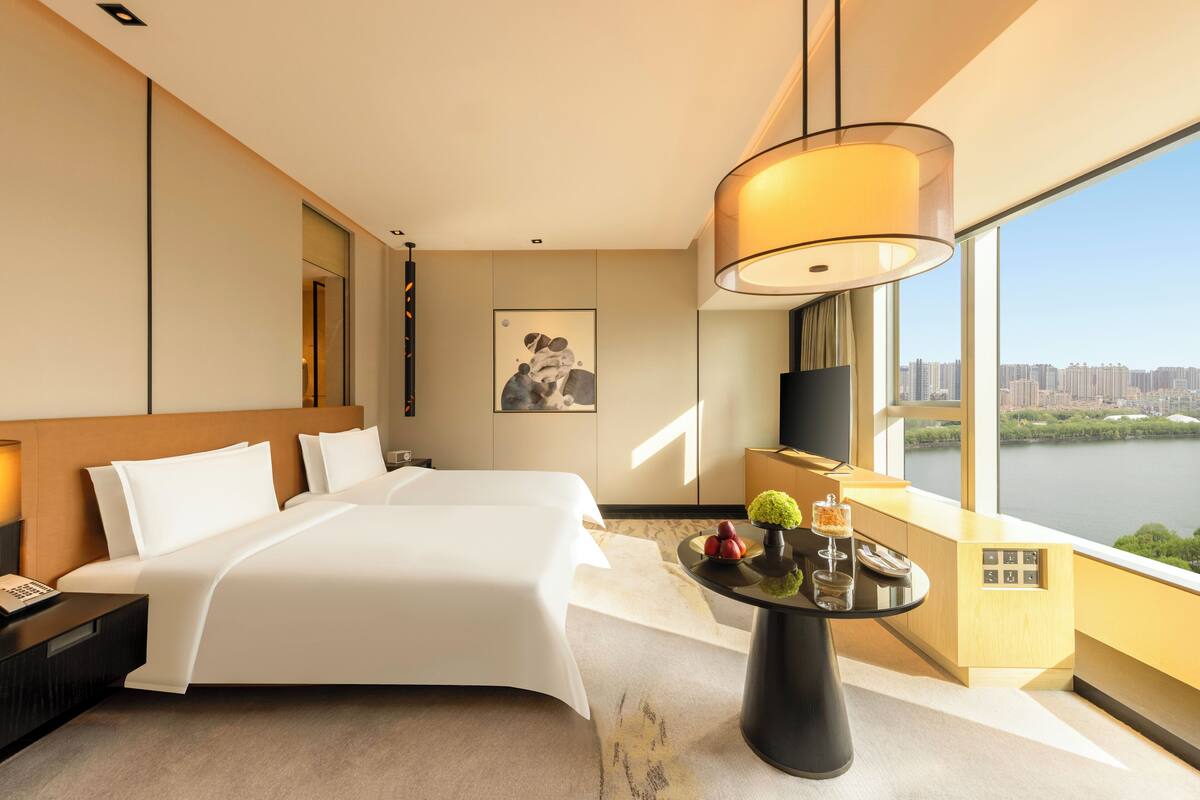 Une chambre d’hôtel avec un grand lit, un bureau avec une lampe, une petite table agrémentée d’objets décoratifs, une télévision et une vue sur une rivière et le paysage urbain.