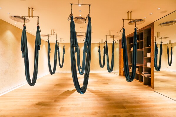 A yoga studio with aerial silks suspended from the ceiling, wooden flooring, and a wooden shelving unit with yoga mats.