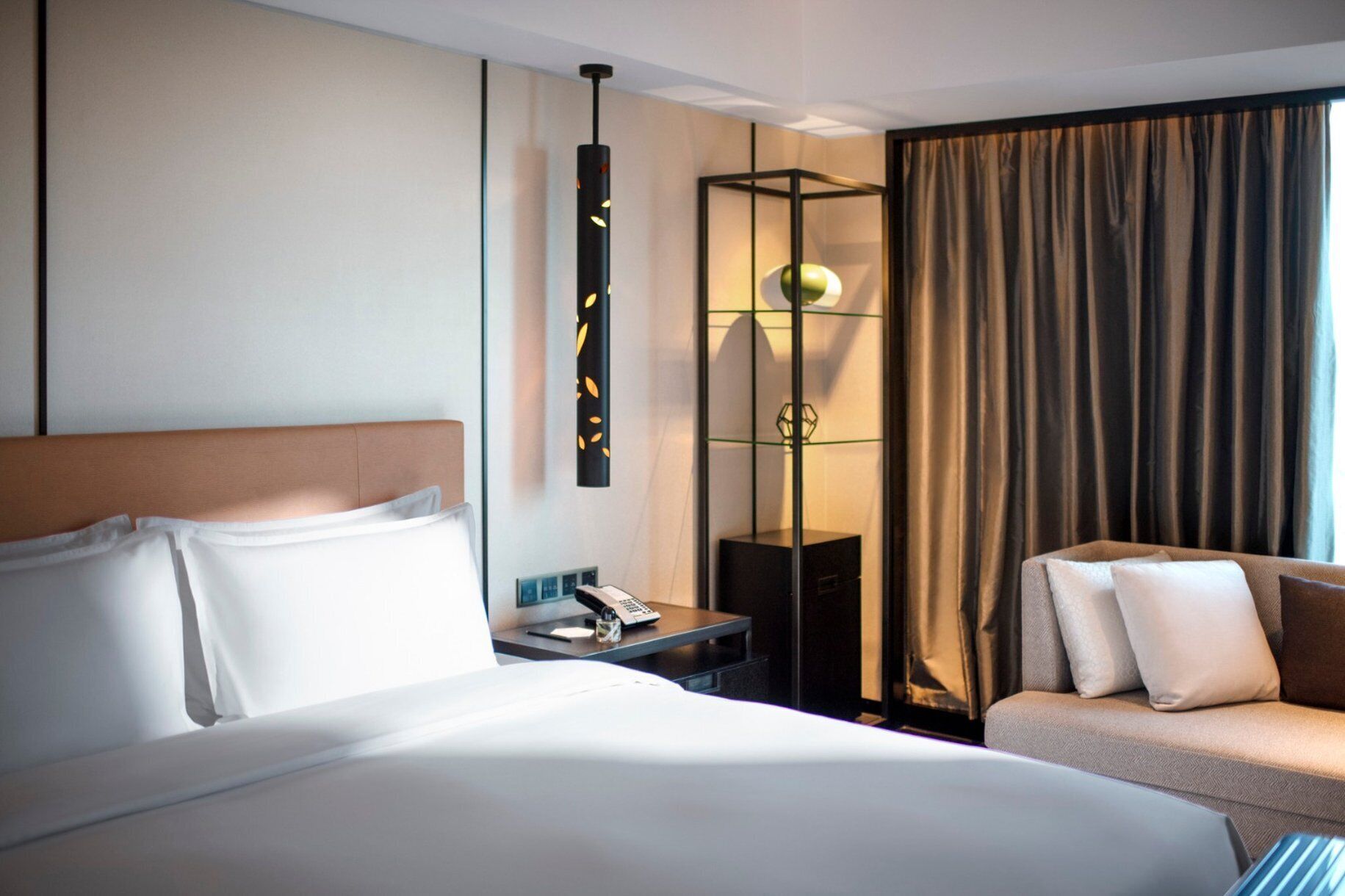 A hotel room with a large bed, a desk with a lamp, a chair, and a view of the city through curtains.