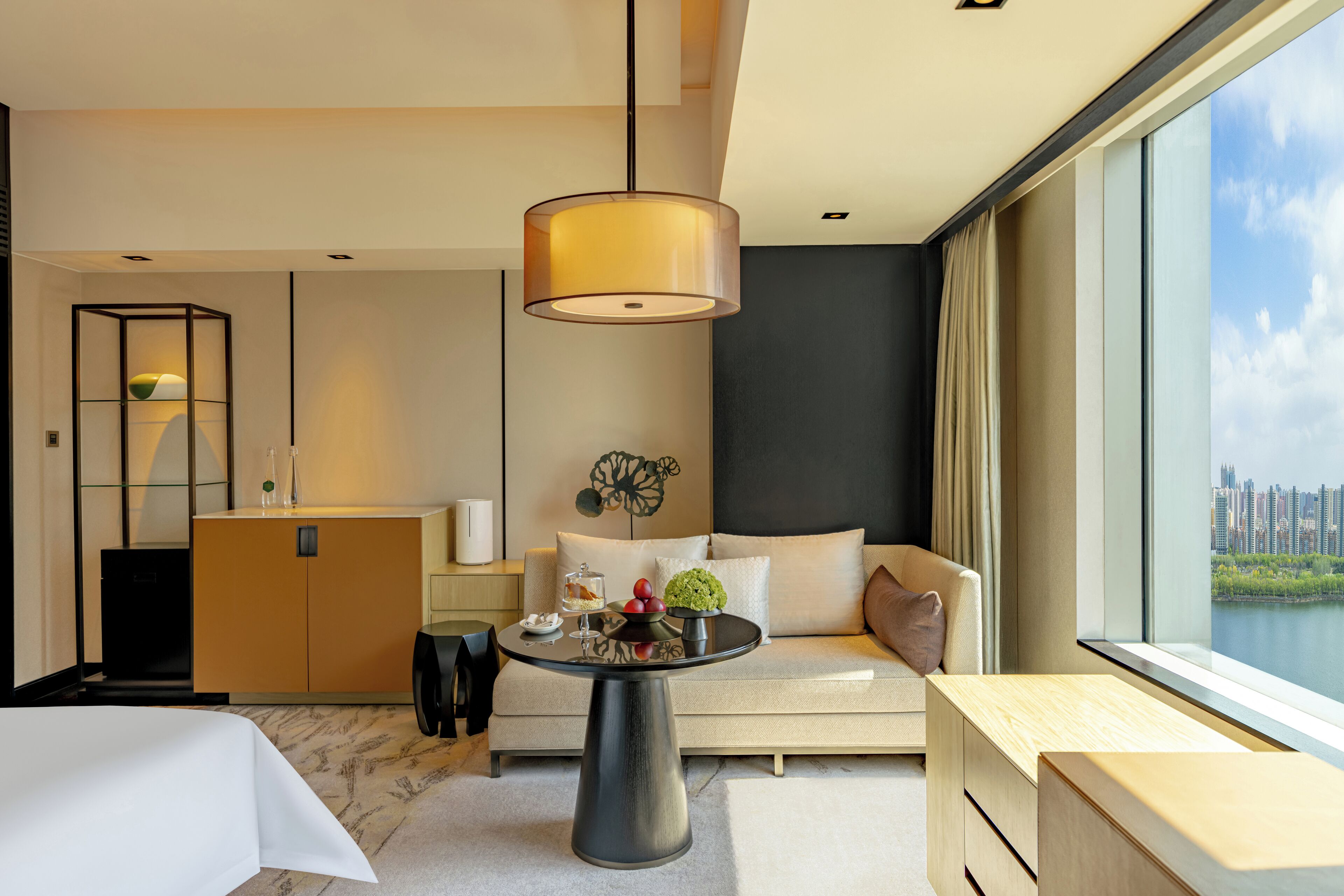 Una habitación de hotel moderna con sofá, una mesa redonda con lámpara, vistas de la ciudad y un ventanal grande.