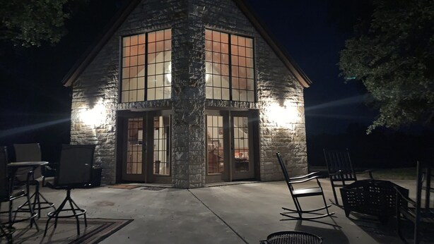A stone building with large windows and a door, outdoor seating, and a grill. - ANTLER OAKS CABIN  (San Saba)