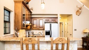 A kitchen with wooden cabinets, a stone island with two chairs, a deer head mounted on the wall, and a refrigerator. - ANTLER OAKS CABIN  (San Saba)