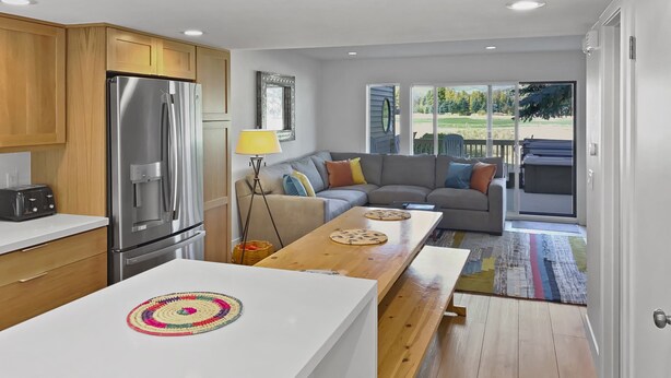 A modern kitchen with a stainless steel refrigerator, wooden cabinets, and a white countertop. There is a dining area with a wooden table and benches, and a gray sofa with colorful cushions. A large window offers a view of a green landscape. - Relax, Recharge and Explore at Vantage 71 (Park City)