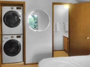Ein Hotelzimmer mit zwei Frontlader-Waschmaschinen von LG, einem runden Spiegel, einer Holztür und einem kleinen Fenster mit Blick auf Bäume.
