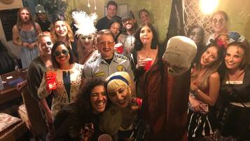 A group of people in a room with a brick wall, a chandelier, and a patterned tapestry.