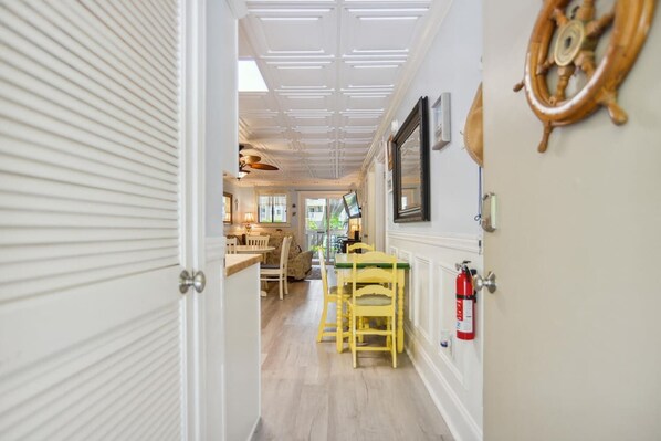 A corridor with a dining area featuring wooden chairs and a table, a fire extinguisher mounted on the wall, and a ceiling with a grid pattern. - Beachside Bliss Villa with Pools & Tennis (Hilton Head Island)