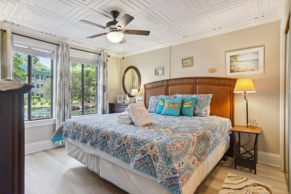 A bedroom with a large bed, a ceiling fan, a nightstand with a lamp, and a view of greenery outside.