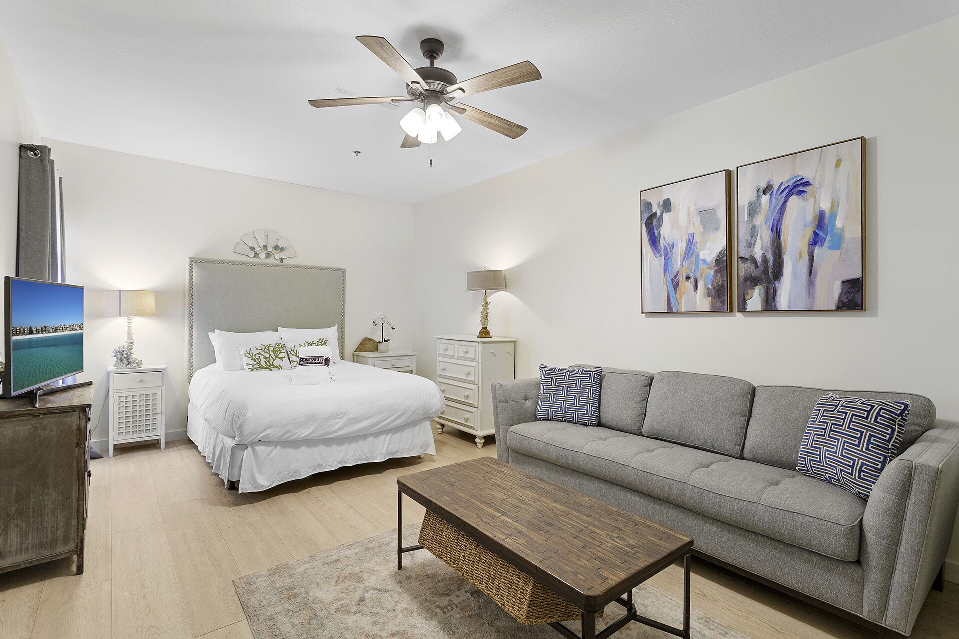A modern hotel room with a bed, a sofa, a coffee table, a TV, and artwork on the walls.