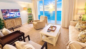 A living room with a television, a sofa, a coffee table, and a balcony with a view of the ocean.