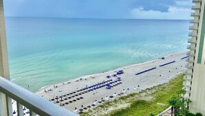 A beach view from a balcony with rows of sun loungers and umbrellas, a clear sky, and the ocean.