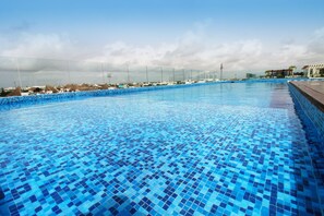 A large, clear blue pool with a tiled floor, surrounded by a fence and overlooking a cityscape with buildings and a cloudy sky.