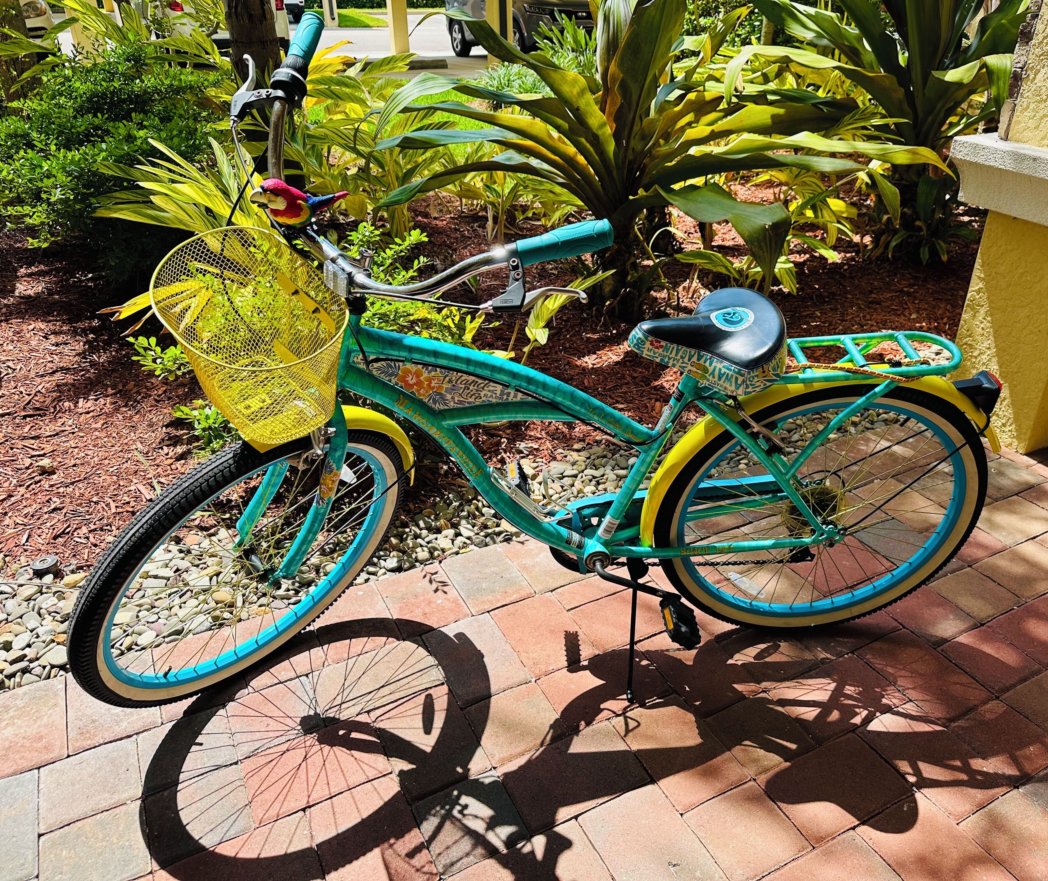 A turquoise bicycle with a yellow basket and a black seat, parked on a tiled patio with plants in the background.