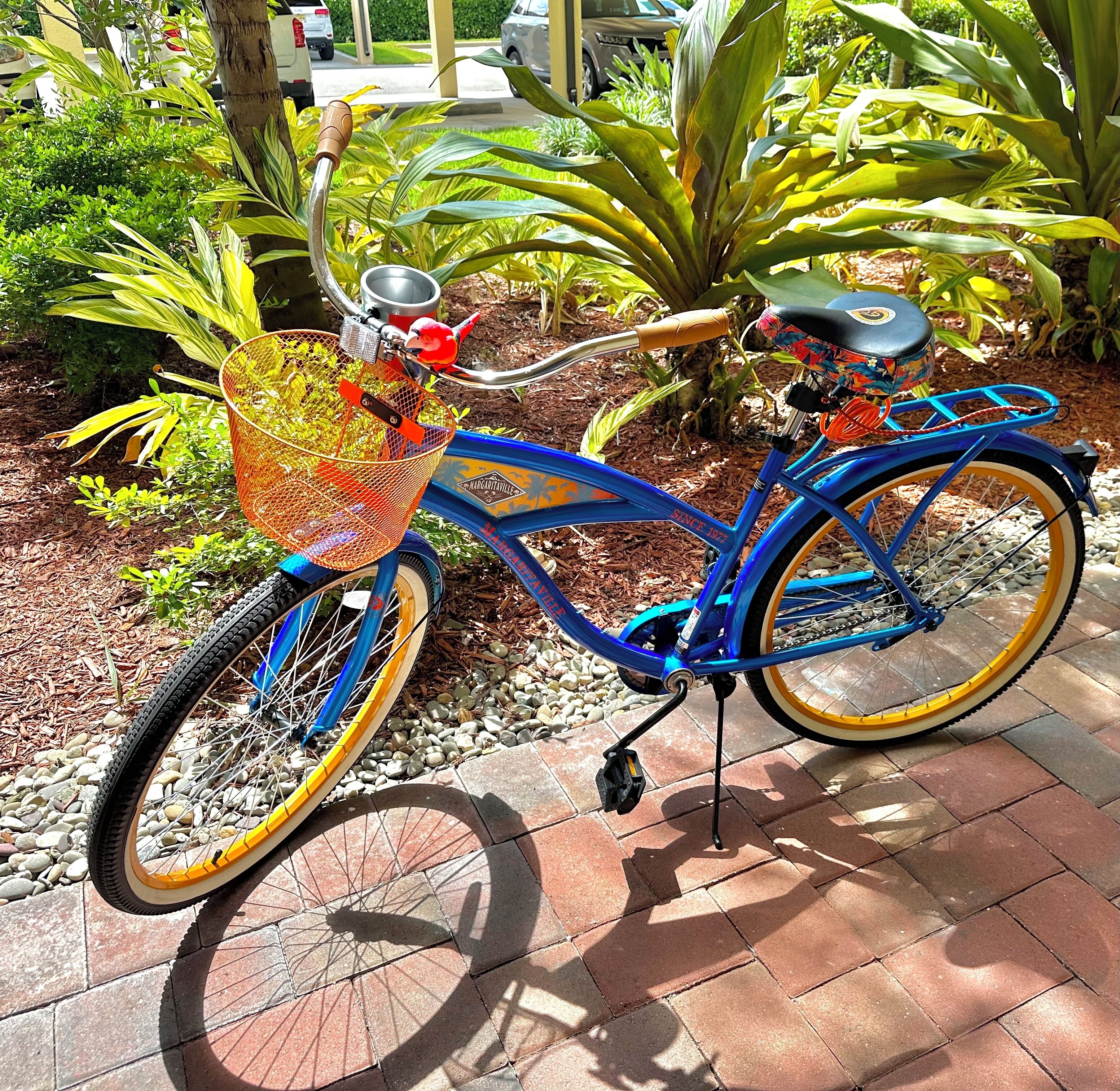 A blue bicycle with a basket and a seat, parked on a paved surface with plants and trees in the background.