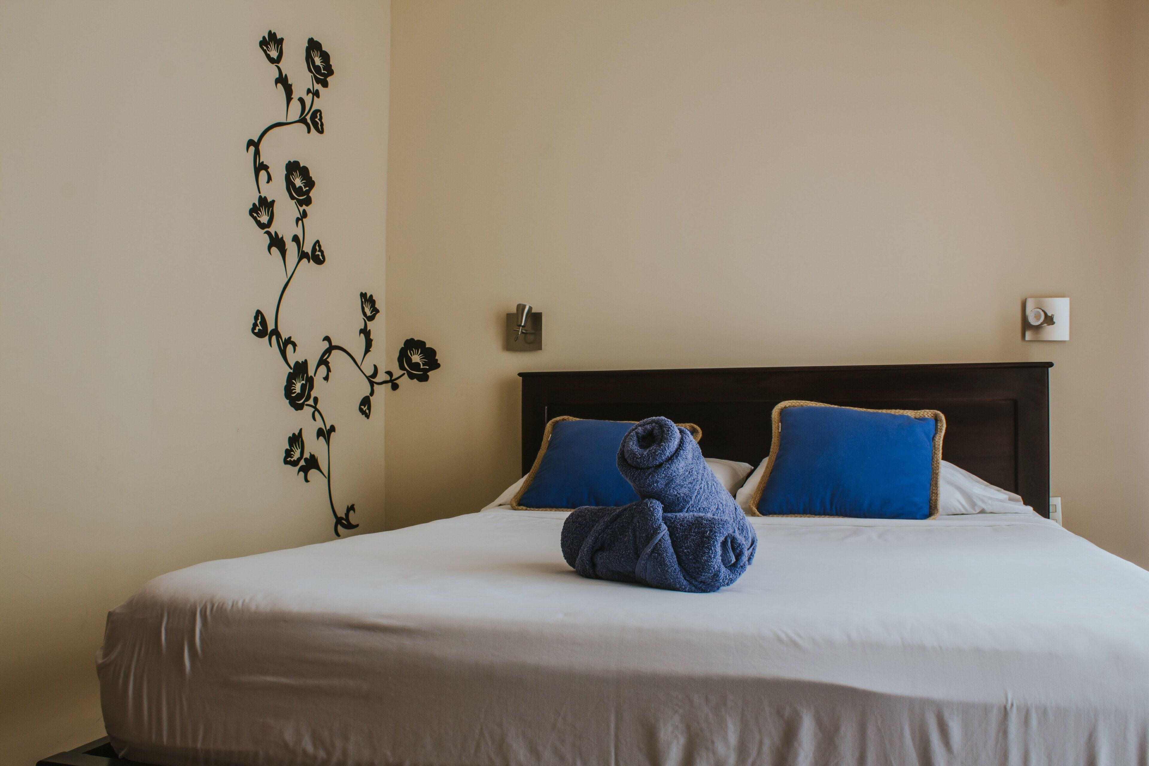 A neatly made bed with blue pillows and a rolled towel, a wall-mounted light switch, and a floral wall decal.