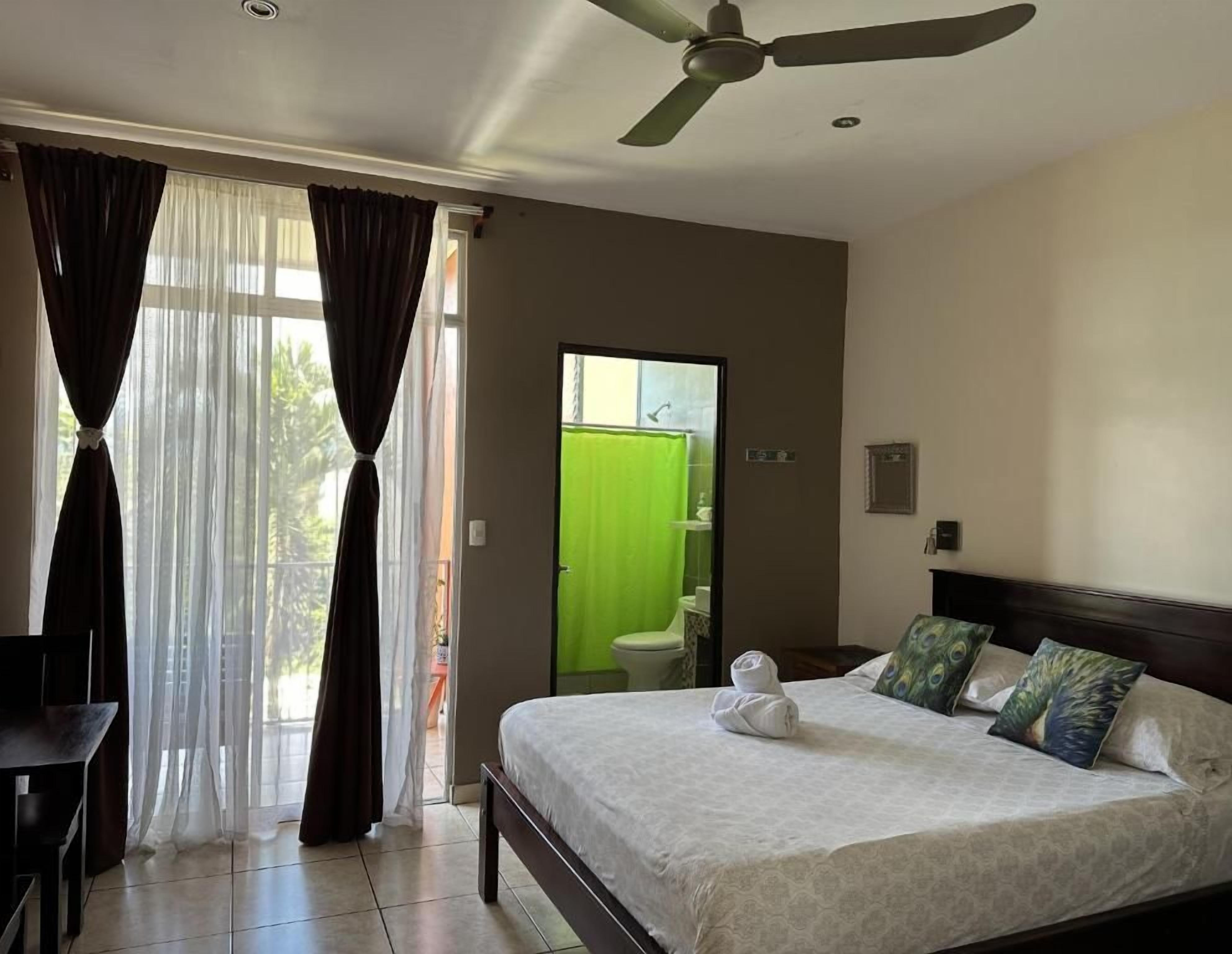 A hotel room with a bed, curtains, a ceiling fan, and a view of the outside.