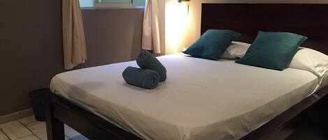 Una cama bien hecha con cabecera, una lámpara de noche y una ventana con cortinas.