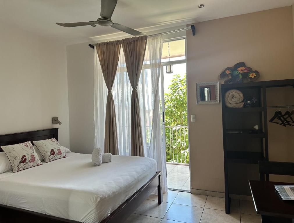 Una habitación de hotel con una cama, un ventilador de techo, una ventana con cortinas, una puerta a un balcón y una pequeña mesa con una silla.