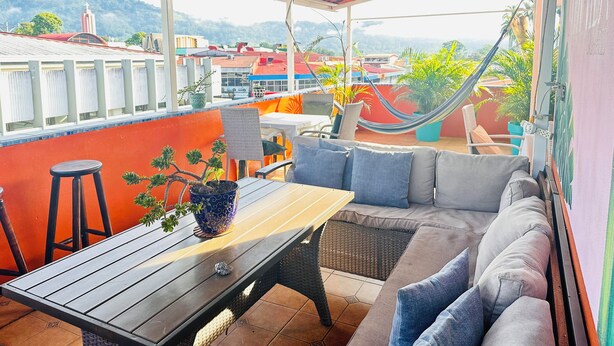 Eine überdachte Terrasse mit Tisch und Sitzgelegenheiten, mit Blick auf eine Stadtlandschaft und dahinter Bergen.