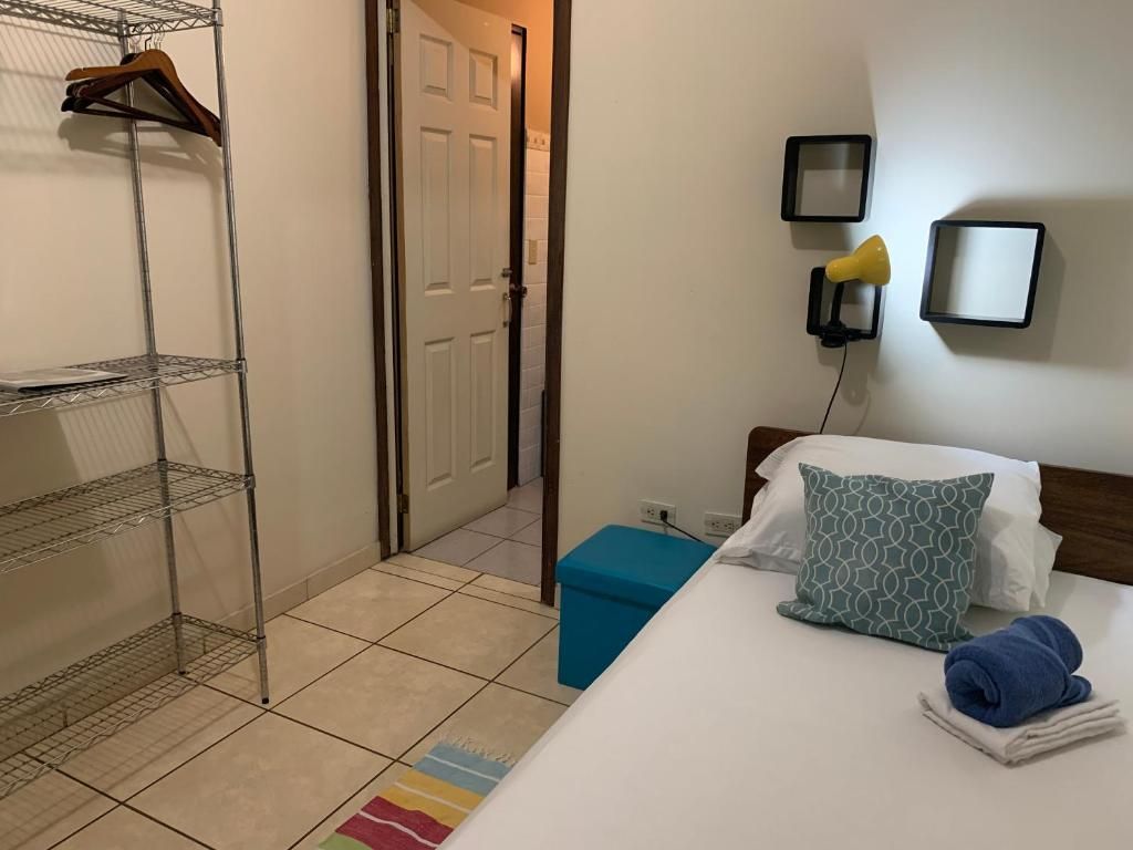 Una habitación de hotel con una cama, un reposapiés azul, un armario con perchas y una puerta que da a otra habitación.