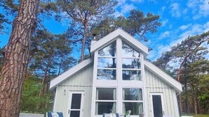 Una cabaña de madera blanca con un amplio ventanal frontal y un balcón, rodeada de árboles.