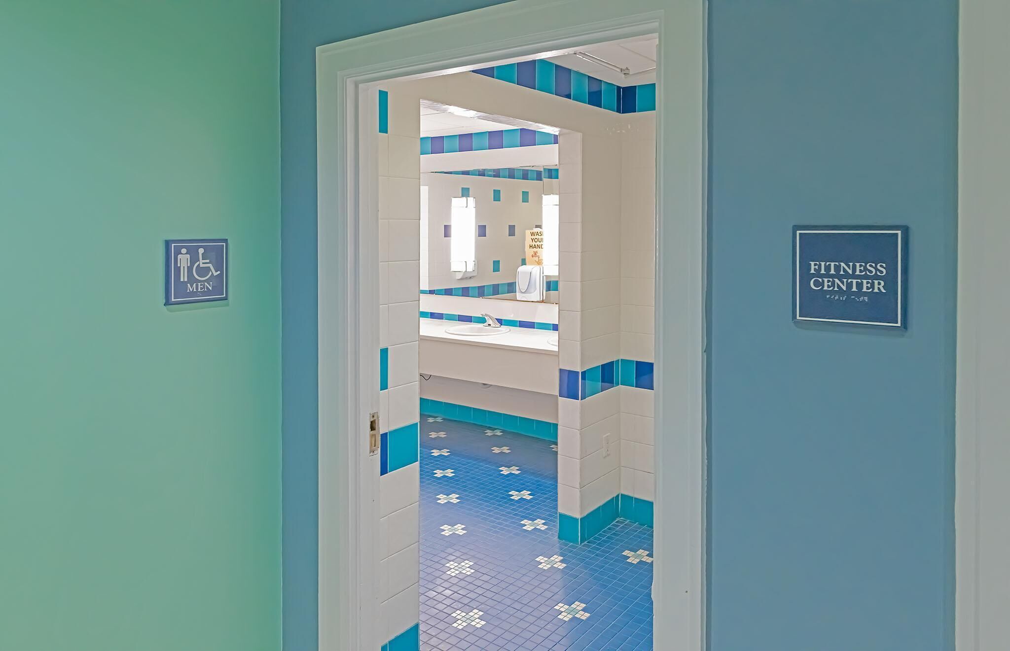 Pasillo que conduce a un gimnasio, con piso de baldosas azules y blancas y un letrero que indica el baño de hombres.