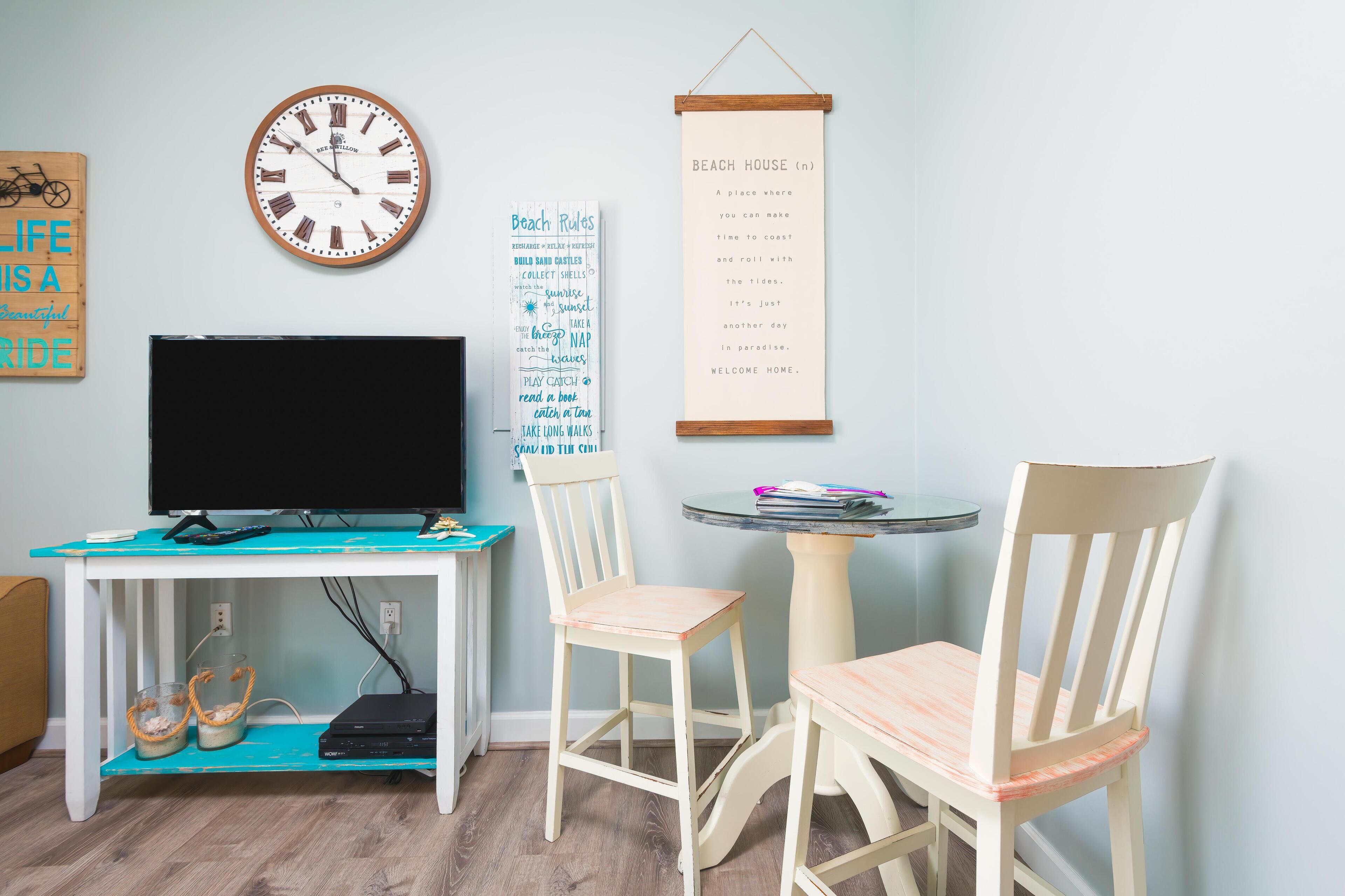 A room with a television, a clock, a wall-mounted sign, and a small table with chairs.