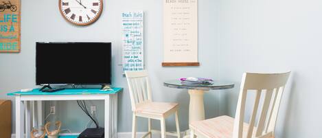 A room with a television, a clock, a wall-mounted sign, and a small table with chairs.