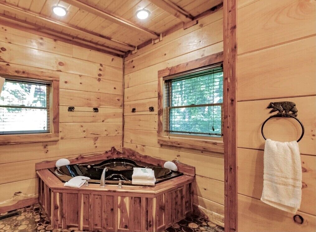 Interior de una cabaña de madera con una bañera de madera, dos ventanas y un perchero para toallas.