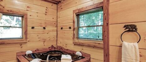 Interior de una cabaña de madera con una bañera de madera, dos ventanas y un perchero para toallas.