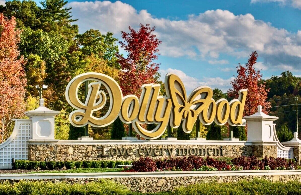 Ein Schild mit "Dollywood" in großen, goldenen Buchstaben, umgeben von einer Steinmauer und viel Grün.