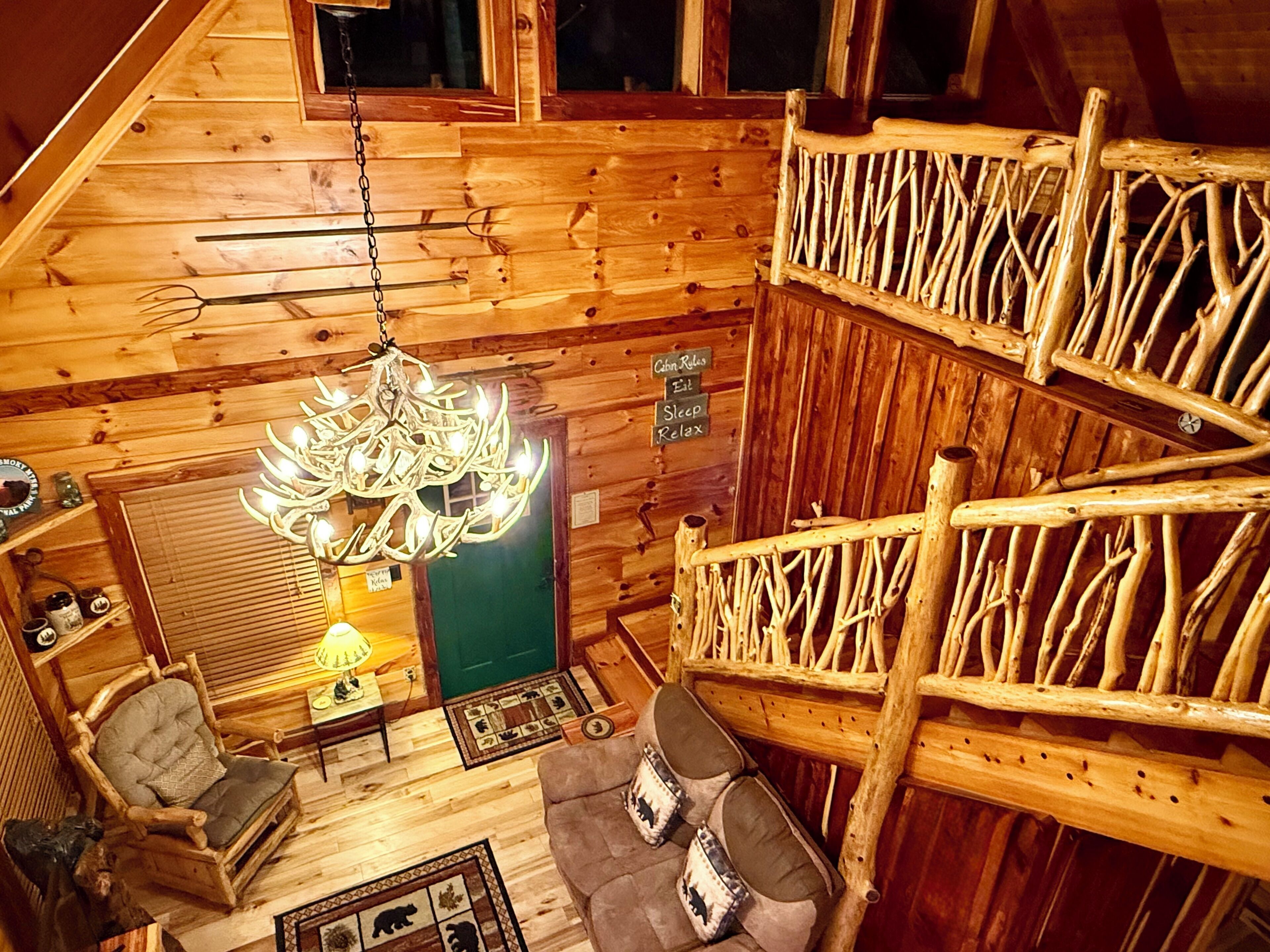 A cabin interior with a wooden ceiling, a chandelier, a couch, a chair, and a wooden railing.