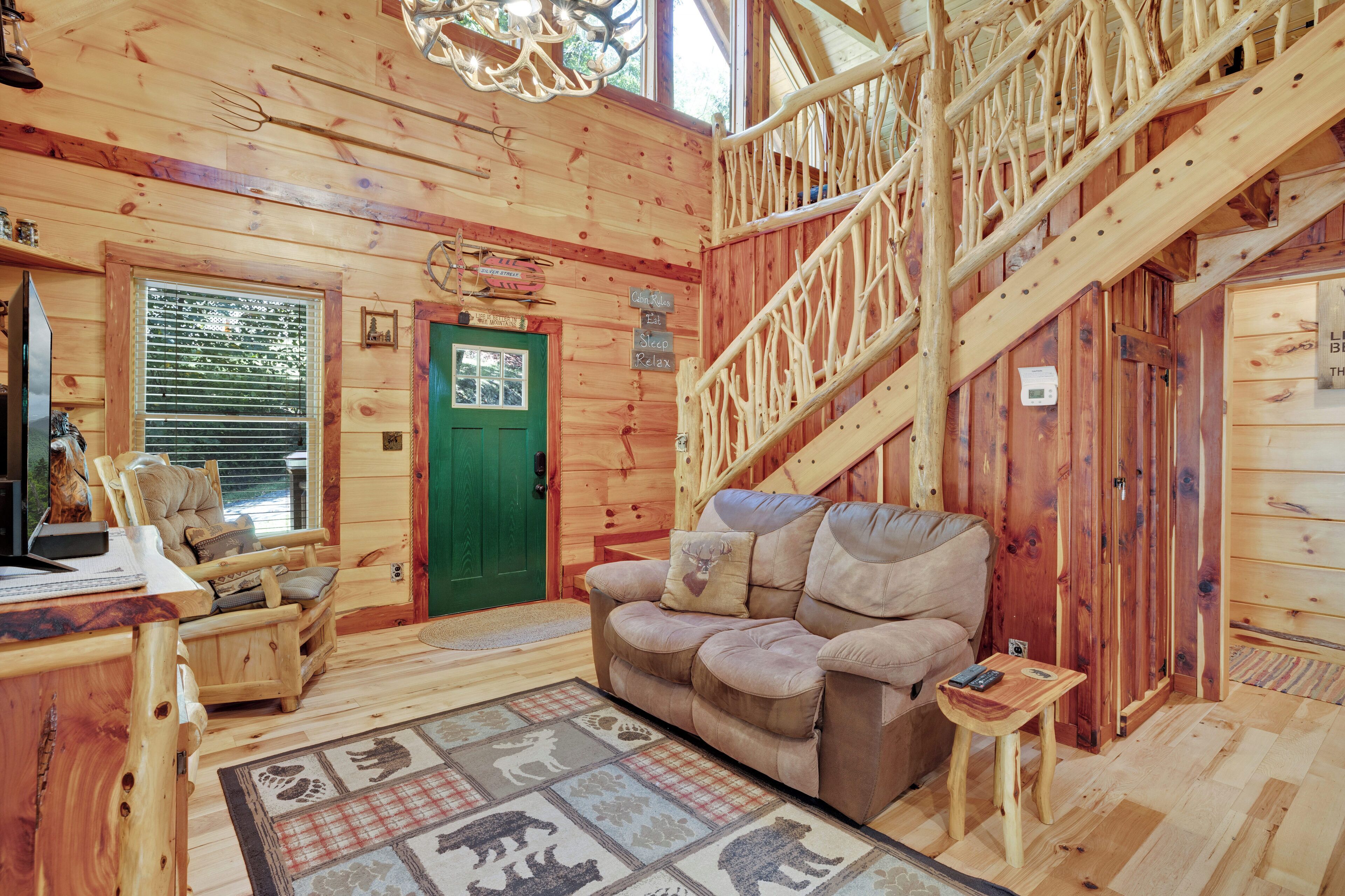 A cozy cabin interior with a wooden staircase, a green door, a sofa, and a desk with a computer.