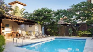 Uma piscina retangular com borda revestida, rodeada por um espaço de terraço com espreguiçadeiras e uma mesa pequena.