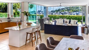 A modern kitchen with a large island, bar stools, and a dining area with a view of the marina. - Pavillions 2 Luxury 4 Bedroom 3 Bathroom With In-ground Pool And Golf Buggy (Whitsundays)
