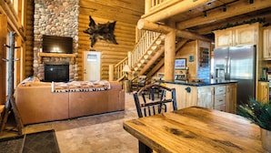 A log cabin interior with a stone fireplace, wooden dining table, and a kitchen area with a refrigerator. - The Lodgepole at Ponds Lodge Cabin 25 (Island Park)